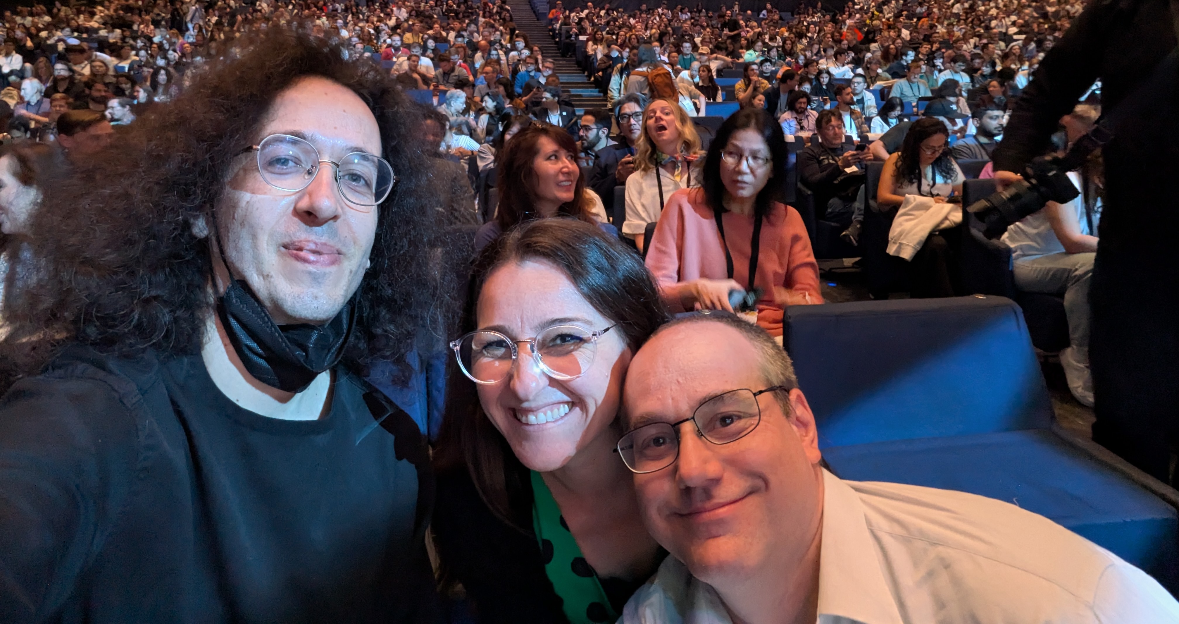 Three smiling faces in front of a large crowd at the auditorium, from left to write: Pedro, Heloisa and Pablo